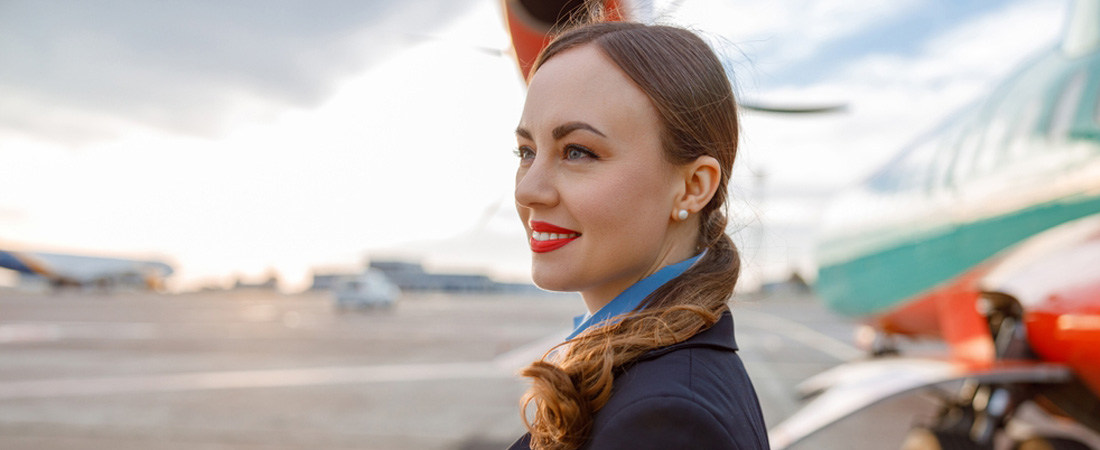 Mujer sonriente con uniforme de tripulante de cabina junto a un avión en la pista de un aeropuerto.